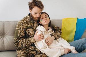 casal ucraniano, militar de uniforme com a namorada no sofá em casa em um fundo de bandeira amarela e azul foto