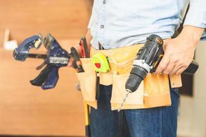 trabalhador manual no cinto de ferramentas e segurando a broca nas mãos, carpinteiro trabalhando com equipamentos na oficina de madeira, homem fazendo marcenaria na carpintaria foto