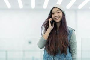 alegre jovem mulher asiática em roupas casuais tendo uma conversa telefônica em pé no shopping, sorrindo freelancer feminina ligando para o colega para compartilhar boas notícias sobre negócios foto