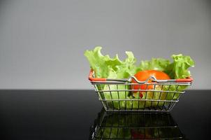 legumes na cesta de compras em uma mesa preta com um reflexo. alface verde e tomate na cesta. foto com espaço de cópia.