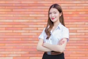 retrato de um estudante tailandês adulto em uniforme de estudante universitário. menina bonita asiática em pé com os braços cruzados sobre um fundo de tijolo. foto