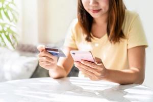 jovem mulher asiática segurando um cartão de crédito e usando telefone inteligente para fazer um pagamento de compras on-line em casa foto