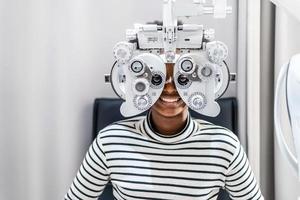 cabelo afro americano africano jovem sorridente fazendo teste de olho no foróptero óptico, verificando seu olho com máquina de optometria foto