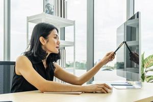 retrato de mulher de negócios asiáticos apontando com caneta e pensando sobre um assunto enquanto está sentado no escritório com seu computador foto