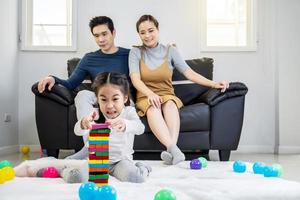 feliz família asiática pai e mãe com garotinha asiática sorrindo brincando com a construção de uma torre de blocos de madeira, pegando telhas por baixo até cair, em momentos bons em casa foto