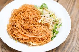 aletria de macarrão frito na chapa branca, macarrão frito estilo chinês com broto de feijão e coentro na mesa de madeira - comida tailandesa foto