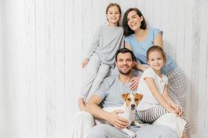 retrato de família feliz interior. lindo pai segura cachorro, linda mãe morena e duas filhas, se divertem juntos, posam para o álbum de família, passam tempo juntos. pessoas, conceito de relacionamentos foto