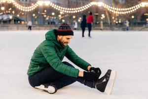 homem de aparência agradável usa casaco verde e chapéu, senta-se no gelo e amarra os patins, vai patinar, tem uma expressão feliz. homem bonito passa férias de inverno no ringue de skate. conceito de inverno e temporada foto