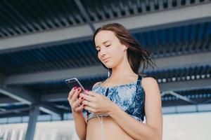 foto de adolescente de aparência agradável com cabelos escuros, segura celular moderno, mensagens de texto em redes sociais, ouve músicas favoritas com fones de ouvido, usa blusa casual, fica ao ar livre. pessoas e conceito de música