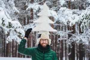 engraçado homem barbudo com barba, vestido com roupas quentes de inverno, segura abeto artificial na cabeça, posa contra árvores cobertas de neve, estando de bom humor. homem otimista passa o tempo ao ar livre foto