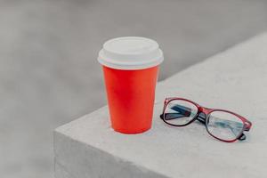 copo descartável vermelho de bebida quente na parede de concreto branco, óculos ópticos próximos. café para viagem em recipiente de papel. ninguém. expresso aromático. foto