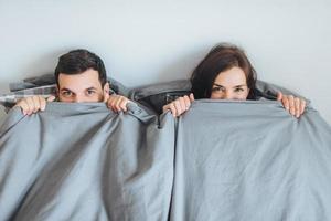 casal alegre amoroso olha do cobertor, deita-se juntos na cama, feliz depois de passar uma noite inesquecível, descansa no quarto. esposa e marido cobertos com roupas de cama, passam a primeira noite de casamento foto