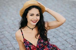 elegante mulher morena bonita com maquiagem usando chapéu de palha na cabeça e vestido de verão sorrindo agradavelmente para a câmera enquanto posava ao ar livre. feliz linda jovem modelando na rua foto