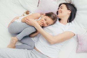 criança do sexo feminino encantada ri alegremente enquanto brinca com a mãe na cama confortável, tem sorrisos positivos nos rostos, usa pijama, tem bom humor após o despertar. pessoas, lazer e conceito de dormir foto