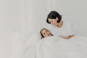 mãe carinhosa brincalhão e sua filha ficam na cama por muito tempo, posam sob o cobertor macio, aproveitam o bom dia, têm sorrisos felizes nos rostos, isolados sobre fundo branco. conceito de maternidade. foto