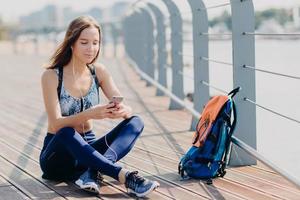 tiro horizontal de bela mulher atlética feliz em tênis, roupas esportivas, sente-se relaxado enquanto se senta ao ar livre, usa telefone celular moderno para navegar na internet, baixa música para lista de reprodução, ouve música foto