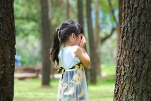 sorridente menina cobrindo os olhos com as duas mãos, brincando de esconde-esconde ao lado de uma grande árvore. menina bonitinha se divertindo ao ar livre no parque.