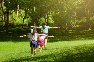 duas filhas adoráveis correndo com a mãe abrindo os braços como se estivessem voando em um jardim de primavera. vista traseira da jovem mãe brincando com crianças no parque. família feliz se divertindo no parque.