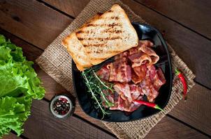 bacon frito e torradas em um prato preto em um fundo de madeira foto