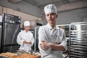 retrato do jovem chef masculino asiático em uniforme de cozinha branco, olhando para a câmera, braços cruzados com confiança, ocupação profissional de alimentos, trabalhos culinários de pastelaria comercial em uma cozinha de restaurante. foto