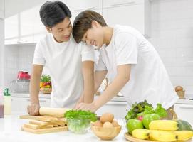 jovem casal lgbt masculino asiático feliz passando tempo juntos durante o cozimento da salada de pão na cozinha branca com um sorriso no rosto. foco seletivo. foto