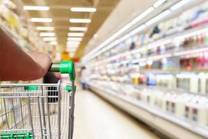 mão de mulher segura carrinho de compras de supermercado com prateleiras de geladeira de desfoque abstrato com garrafas de leite fresco e produtos lácteos na mercearia foto