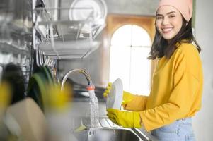 jovem mulher feliz usando luvas amarelas lavando pratos na cozinha em casa foto