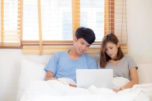 lindo jovem casal asiático sorri navegando na internet assistindo filme online juntos na cama com o computador portátil para relaxar juntos, mulher asiática e homem trabalham notebook no quarto, conceito de estilo de vida. foto