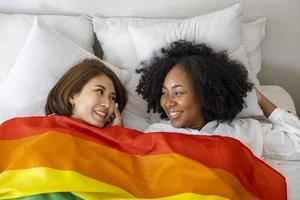 casal do mesmo sexo casamento de raças diferentes com bandeira lgbtq arco-íris para o mês do orgulho enquanto deitados juntos na cama para promover a igualdade e as diferenças do conceito homossexual foto