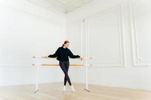 dançarina de balé clássico tem treino de alongamento, fica perto de barras de balé, usa moletom preto e leggings, faz exercícios em estúdio, focado. instrutora certificada de coreografia foto