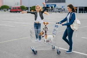 conceito de compras e família. feliz mulher carrega carrinho em que garotinha fica com cachorros, posar contra o fundo do shopping, alegrar novas compras. pessoas, felicidade, conceito de dia de folga foto