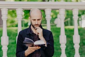 macho careca barbudo sério focado segura o queixo, lê atentamente o livro, posa ao ar livre contra a varanda branca, concentrado na trama, gosta de tempo livre e dia ensolarado de verão ao ar livre. descansar no parque. foto