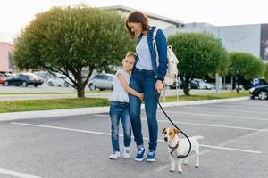 mãe afetuosa abraça filha pequena, passear ao ar livre junto com seu animal de estimação favorito, aproveitar o bom dia e o clima, posar contra o fundo da cidade. família, estilo urbano e conceito de dia de folga foto