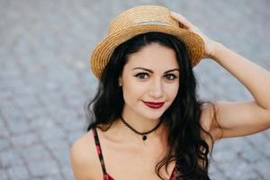 retrato ao ar livre da mulher morena com olhos escuros, lábios vermelhos e pele saudável usando chapéu de verão, mantendo a mão na cabeça, descansando enquanto caminhava na rua da cidade antiga. juventude, conceito de moda foto