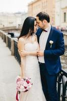 casal romântico fica na ponte fica perto um do outro, olha carinhosamente um para o outro, sente amor e paixão. lindo noivo e noiva em roupas de casamento expressam seus sentimentos. foto