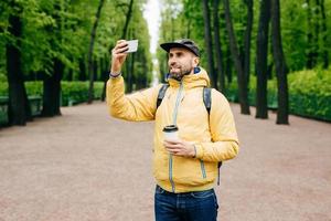 retrato ao ar livre de bonitão com barba grossa vestindo amarelo anoraque e jeans segurando mochila, café e smartphone fazendo selfie contra árvores verdes. turista satisfeito descansando no parque foto