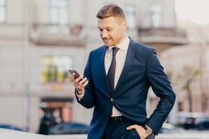 foto de empresário masculino de sucesso segura telefone inteligente moderno nas mãos, lê notícias da troca financeira, navega na internet, faz pagamentos on-line, tem pausa no trabalho, vai a um café ou restaurante