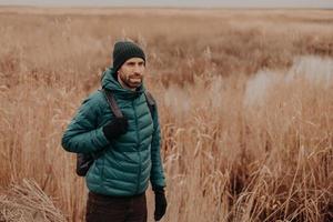 tiro ao ar livre de viajante masculino pensativo vestido com roupas quentes, de bom humor, tem mochila nas costas, caminha em ambiente rural perto de campo durante o outono, goza de uma atmosfera calma. conceito de lazer foto