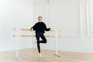 mulher magra positiva tem aula de dança, pratica no estúdio perto da barra de balé, fica em uma perna, usa roupa preta e sapatos esportivos brancos, tem músculos fortes, forma corporal agradável. conceito de ginástica foto