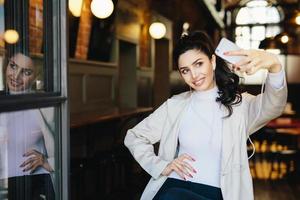 mulher morena alegre em roupas elegantes brancas segurando o telefone móvel genérico fazendo sefie contra o fundo do café aconchegante. elegante jovem feminina fazendo foto de si mesma se divertindo. tecnologia moderna