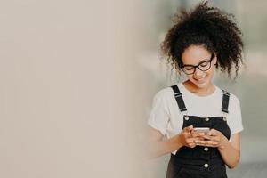linda garota afro-americana em roupas da moda, usa o celular para enviar mensagens de texto, conectado ao wifi, verifica a caixa de e-mail, fica perto da parede branca com espaço vazio. comunicação online foto