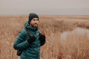 cara com a barba pensativa usa chapéu, jaqueta verde e luvas, carrega mochila, posa contra o fundo amarelo do campo de outono, passeia lá fora, respira ar fresco, focado na distância. conceito de turismo foto