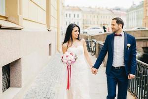 mulher atraente com cabelos escuros, usa vestido branco, segura buquê, mantém a mão dos noivos, olha um para o outro com muito amor, anda pela cidade antiga na ponte. conceito de emoções positivas foto