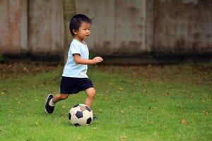 menino asiático jogando futebol no parque. garoto driblando a bola no campo de grama. foto