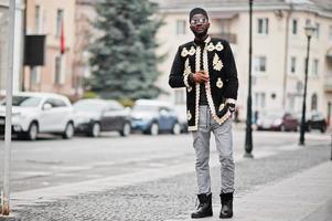 mega homem africano elegante em pose de jaqueta tradicional. elegante negro de chapéu e óculos de sol com charuto na mão. foto