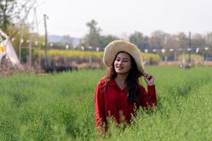 mulher asiática atraente na camisa vermelha com chapéu de agricultor no jardim de plantas de folha verde olhando para a frente com um sorriso feliz foto