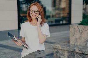 jovem ruiva sorridente empresária em óculos falando no celular ao ar livre foto