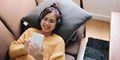 jovem preguiçosa passa o fim de semana em casa deitada no sofá usando fones de ouvido olhando para a tela do telefone inteligente ouvindo música online. foto