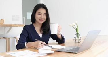 sorridente jovem empresária asiática segurando uma caneca de café e laptop no escritório. olhando para a câmera. foto