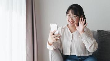 jovem mulher asiática usando smartphone para videoconferência online acenando a mão fazendo gesto de olá no sofá da sala de estar. foto